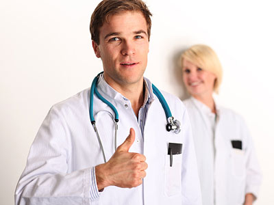 Ein junger Arzt zeigt ein Daumen-hoch-Zeichen, während eine Ärztin im Hintergrund lächelt. Beide tragen weiße Ärztekittel.